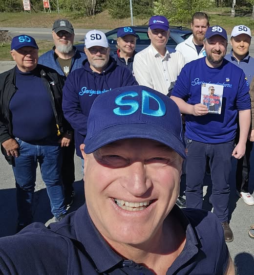 En grupp glad medlemmar från Sverigedemokraterna Vallentuna poserar utomhus i solsken. De bär profilkläder som blå och vita kepsar med "SD"-logotyp samt blå tröjor. En person i förgrunden tar en selfie och en annan person håller upp ett flygblad med texten "Sverige på rätt väg".