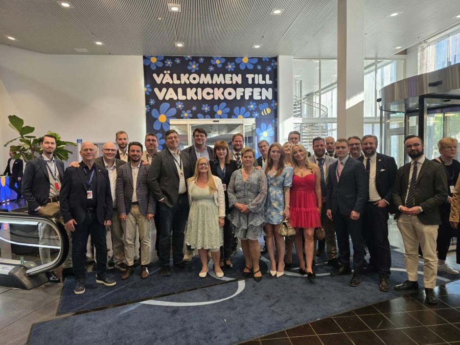 Gruppbild från en entréhall där ett tjugotal personer står samlade på en mörkblå matta. Flera bär ID-brickor runt halsen. I bakgrunden syns glaspartier, en rulltrappa till vänster och en stor blå fondvägg med texten "Välkommen till valkickoffen" dekorerad med Sverigedemokraternas blåsippor och en liten logotyp.