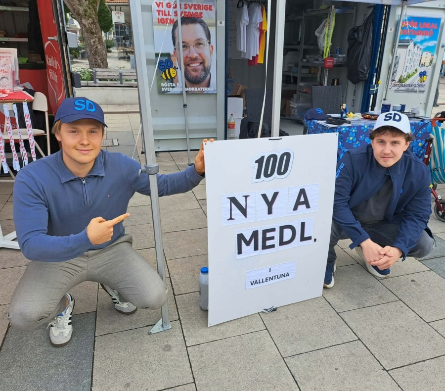 Två unga män i SD-kepsar hukar bredvid en skylt där det står ”100 NYA MEDL. i VALLENTUNA” framför partiets valstuga.