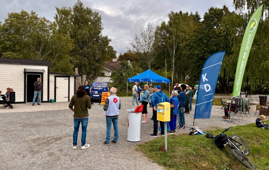 Utomhusmöte på Kårsta Torg under Politiklördag där representanter från flera politiska partier samlats inför valet. I förgrunden syns besökare och partipolitiker vid partitält, valflaggor för KD och MP samt en valstuga.