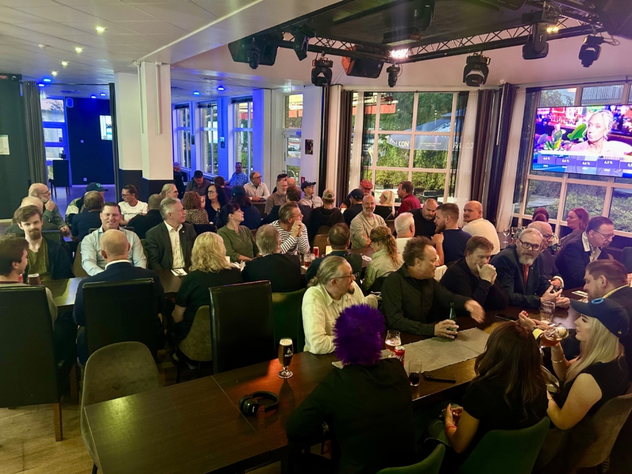 Ett stort antal deltagare samlade vid långbord på Gästis Kök & Bar under SD Vallentunas valvaka, där de följer valresultaten från en tv-skärm på väggen.